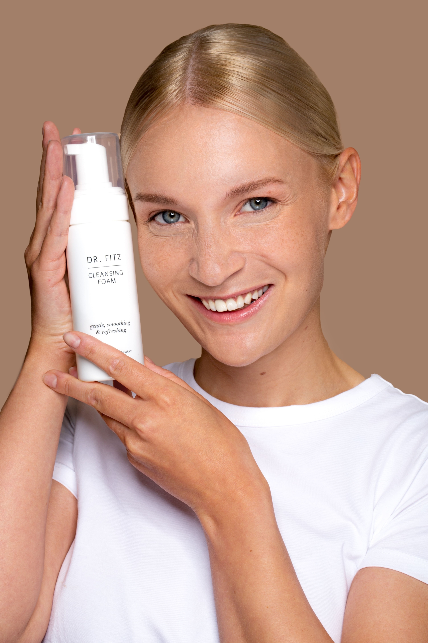 Junge Frau, blond, hellhäutig, blauäugig, schmiegt sich das Produkt Cleansing Foam an ihre Wange und lächelt.